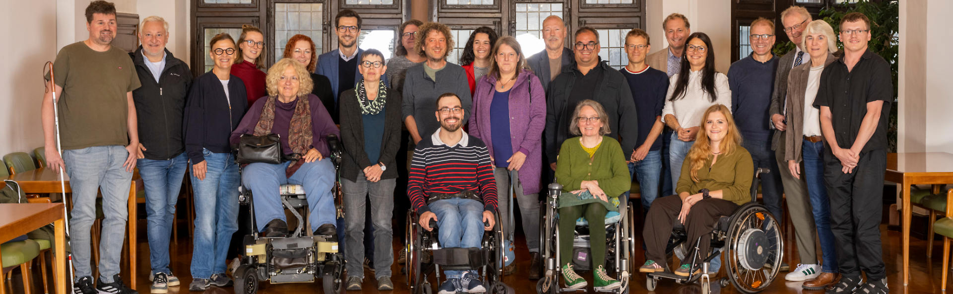 25-bhbr-gruppe2 Gruppenfoto von etwa 25 Personen in einem hellen Raum. Einige Personen stehen, andere sitzen in Rollstühlen oder nutzen Gehhilfen. Alle blicken lächelnd in die Kamera.